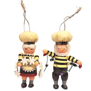 May include: Two Hershey's Christmas ornaments. One is a woman wearing a chef's hat, glasses, and a black and yellow striped outfit. The other is a man wearing a chef's hat, glasses, and a black and yellow striped outfit, holding Christmas lights.