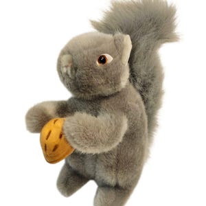 May include: A plush squirrel toy made of soft grey fur with a bushy tail, dark brown eyes, and a small pink nose. The squirrel is holding a golden-yellow plush acorn with brown stitched details between its front paws in an upright position.