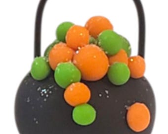 May include: A black cauldron-shaped decoration with a black handle, filled with orange and green round candies. The cauldron has a glittery texture. This Halloween-themed item is perfect for festive decor.