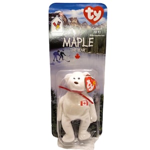 May include: A white plush bear toy named "Maple" with a red ribbon and a Canadian flag patch. The toy is in a clear plastic package with the text "Collect All 47" and "Maple - The Bear".