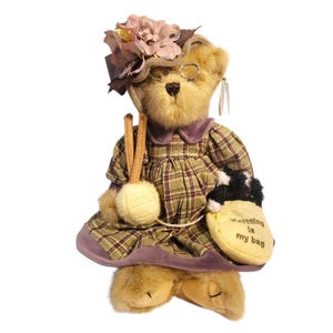 May include: A plush teddy bear wearing a plaid dress, hat with a flower, and glasses. The bear holds a ball of yarn and a bag that says "Knitting is my bag". The bear is a light brown color.