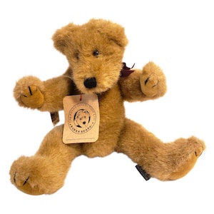 May include: A plush, light brown teddy bear with a tag from The Boyds Collection Ltd. The bear has a black nose, dark brown eyes, and a burgundy ribbon. The bear is in a playful pose with its arms and legs spread.