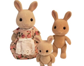 May include: Three tan rabbit figurines. One rabbit wears a pink floral dress with a white apron. The other two rabbits are standing upright. The figurines have black eyes and are designed to look like a family.