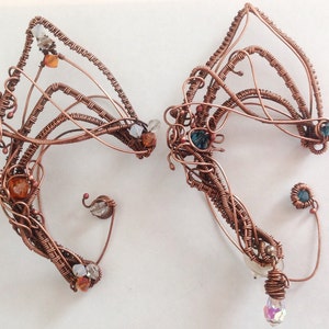 May include: A pair of copper wire ear cuffs featuring a variety of coloured beads and crystals. The ear cuffs are intricately designed with a delicate, organic shape.
