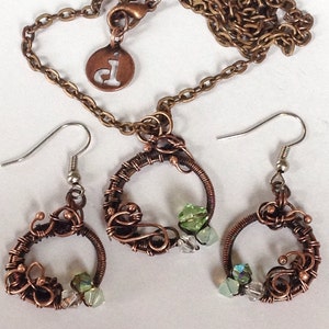 May include: A copper wire necklace and earrings set with green and clear glass beads. The necklace has a circular pendant with a delicate wire design. The earrings are also circular with a similar wire design.