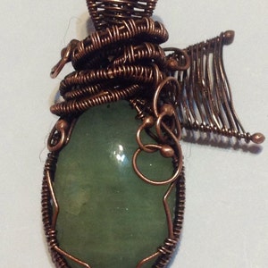 May include: A green gemstone pendant wrapped in copper wire. The wire is woven into an intricate design with a small wing on one side.