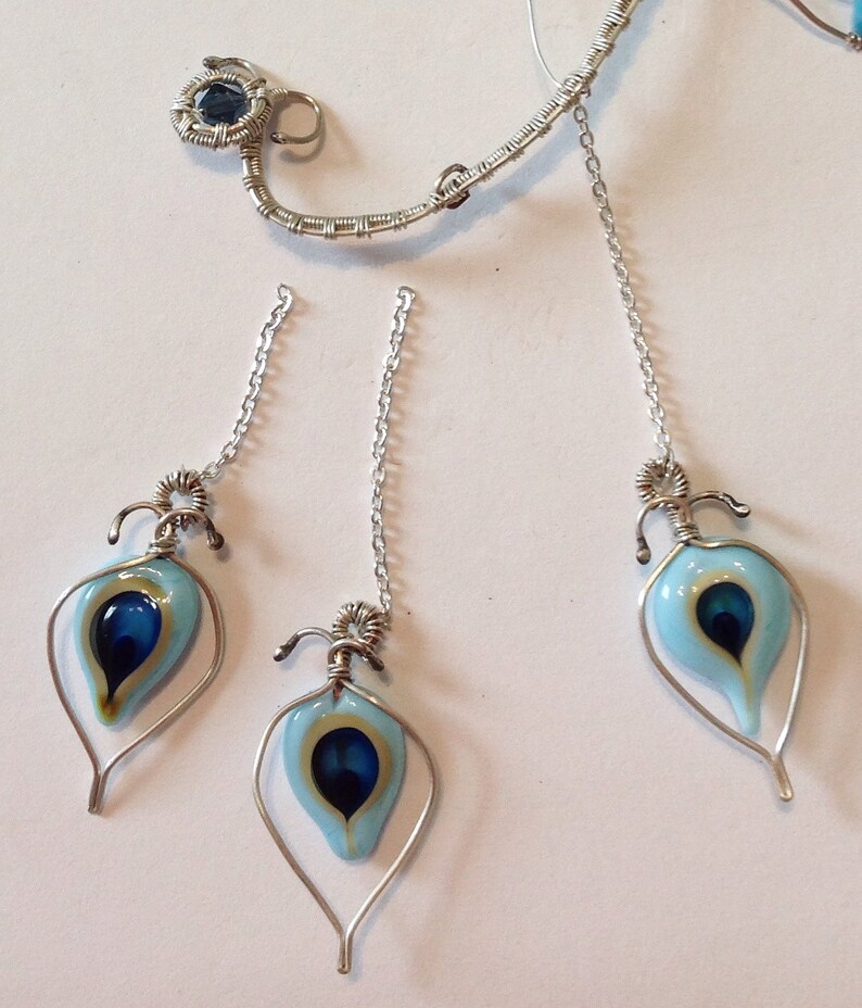 Pu&ograve; includere: Tre ciondoli avvolti in filo d'argento con forme a goccia di vetro blu e oro. Ogni ciondolo &egrave; appeso a una catena d'argento.