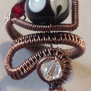 May include: A copper wire ring featuring a black and white swirl glass bead, a red glass bead, a clear crystal bead, and a copper ball.