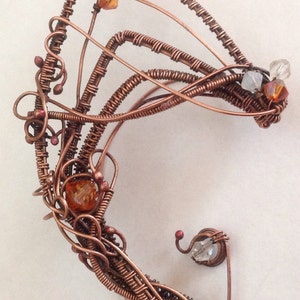 Pode incluir: Um brinco de orelha de fio de cobre com um design delicado e intrincado. O brinco é adornado com pequenos cristais transparentes e âmbar.