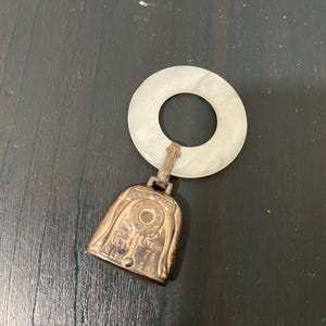 May include: A vintage bell-shaped pendant with a white circular handle. The bell is bronze-colored with engraved text, including "OCT 74" and "4.0z". The handle is a translucent white color.