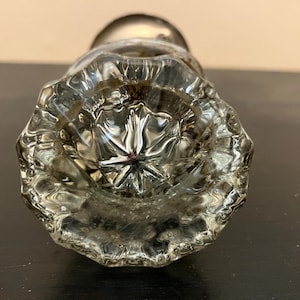 May include: A clear glass door knob featuring a multi-faceted, starburst-like pattern radiating from the center. The knob has a circular, scalloped edge design and a metallic base component visible at the top, reflecting light through the glass.