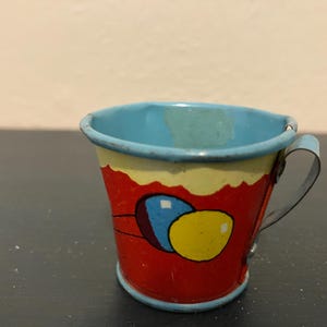 May include: A small, vintage tin cup with a red base, yellow and blue accents, and a light blue interior. The cup features a painted design of balloons and a metal handle. The cup is likely a child's toy or collectible item.