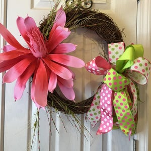 May include: A pink and green wreath featuring a large pink flower and a polka dot bow. The wreath is made from twigs and has a natural look.