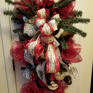 Puede incluir: Una corona de Navidad roja y blanca con un lazo de malla roja y una cinta blanca con el texto "Feliz Navidad" y "Felices Fiestas".