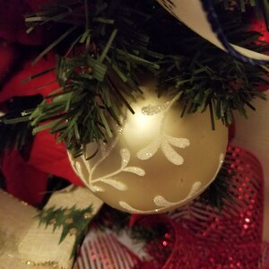 Puede incluir: Un adorno navide&ntilde;o dorado con un dise&ntilde;o de copo de nieve blanco, rodeado de cinta a rayas rojas y blancas y ramas de pino verdes.