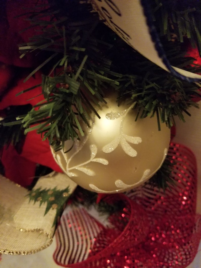 Puede incluir: Un adorno navide&ntilde;o dorado con un dise&ntilde;o de copo de nieve blanco, rodeado de cinta a rayas rojas y blancas y ramas de pino verdes.