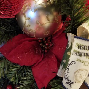 Puede incluir: Una corona de Navidad con una flor de pascua roja, un adorno plateado con dise&ntilde;os de hojas doradas y una cinta de arpillera con el texto "Oh Christmas Tree" impreso en ella.