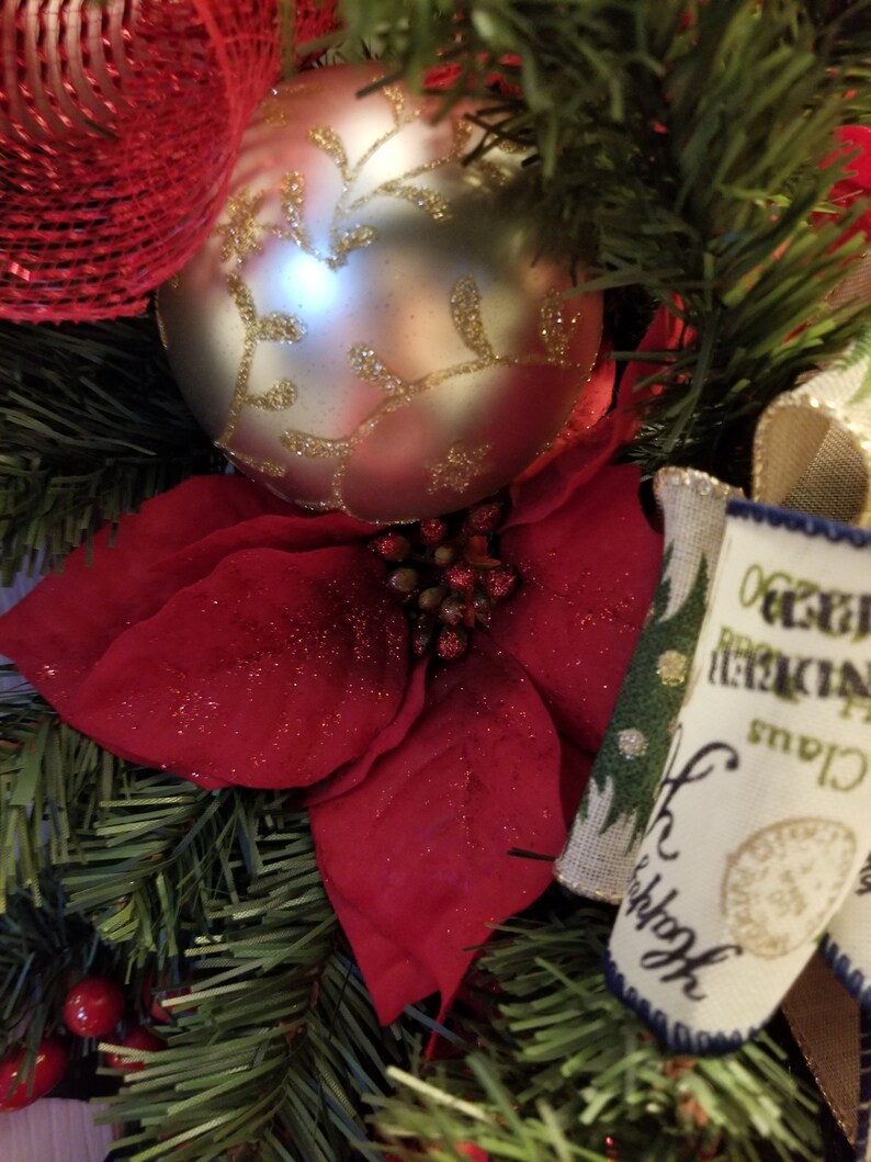 Puede incluir: Una corona de Navidad con una flor de pascua roja, un adorno plateado con dise&ntilde;os de hojas doradas y una cinta de arpillera con el texto "Oh Christmas Tree" impreso en ella.