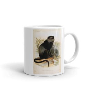 Peut inclure: Une tasse en céramique blanche avec une illustration en noir et blanc d'un singe assis sur une branche. Le singe a un visage noir et une barbe blanche. La tasse a une poignée noire.