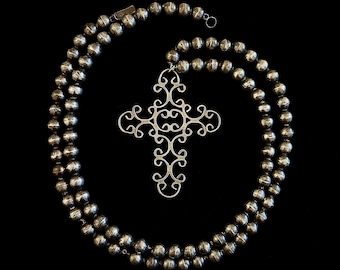 May include: A silver necklace with a decorative cross pendant. The necklace has two strands of round beads. The cross pendant features an ornate, openwork design. The background is solid black, highlighting the jewellery.