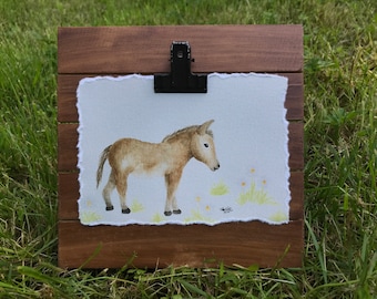 Könnte beinhalten: Aquarellmalerei eines braunen und weißen Fohlens, das auf einer grünen Wiese mit gelben Blumen steht. Das Bild ist in einem Holzrahmen gerahmt.