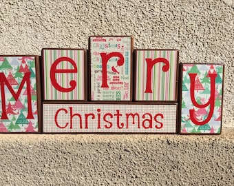 May include: Wooden blocks spelling out "Merry Christmas" in red letters. Each block has a different patterned paper covering. The "Merry" blocks have green and pink stripes, the "Christmas" block has white polka dots, and the "Y" blocks have a pattern of green and pink Christmas trees.