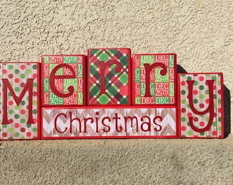 May include: Wooden block letters spelling "Merry Christmas" with red and green plaid, polka dot, and chevron patterns. The letters are stacked on top of each other.