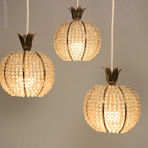 Op de afbeelding: Drie hanglampen met bolvormige kappen van heldere, gefacetteerde kralen. Elke lamp heeft een bronskleurige kroonvormige top. De lampen hangen aan witte snoeren tegen een neutrale grijze achtergrond. De lichten zijn verlicht.