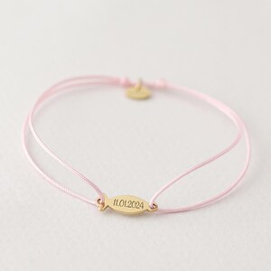 May include: A pink string bracelet with a gold fish charm engraved with the date 11.01.2024.