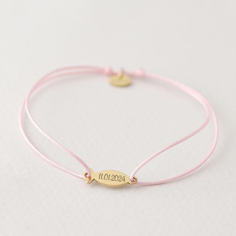 May include: A pink string bracelet with a gold fish charm engraved with the date 11.01.2024.