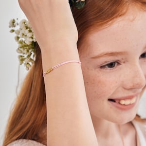 May include: A pink string bracelet with a small gold charm. The charm has the word "love" engraved on it.