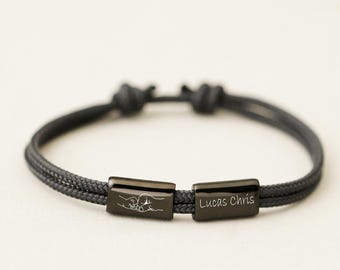 May include: A black cord bracelet featuring two rectangular metal beads. One bead displays an engraved line drawing of two fists bumping, while the other bead is engraved with the text Lucas Chris in a clean, white sans-serif font.