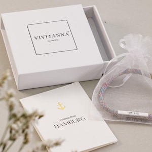 May include: A white box with the text "VIVIANA HAMBURG" on the top. Inside the box is a white card with a gold anchor and the text "Greetings from HAMBURG". A white organza bag with a colorful bracelet inside.