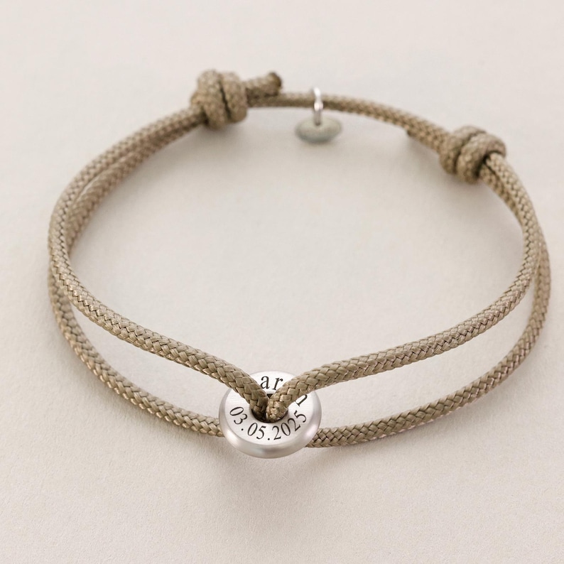 May include: A beige rope bracelet with a silver charm that says "03.05.2025" on it.