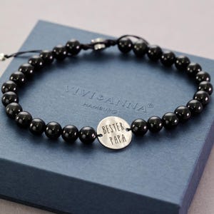 Puede incluir: Pulsera de cuentas negras con un dije plateado que dice "Bester Papa". La pulsera está en una caja azul con el texto "Vivi & Anna Hamburg" en ella.