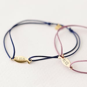 May include: Two personalized friendship bracelets with gold fish charms. One bracelet has a blue cord and the charm says "02.08" with a sun symbol. The other bracelet has a pink cord and the charm says "Alice".