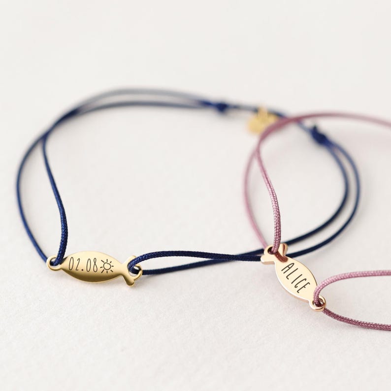 May include: Two personalized friendship bracelets with gold fish charms. One bracelet has a blue cord and the charm says "02.08" with a sun symbol. The other bracelet has a pink cord and the charm says "Alice".