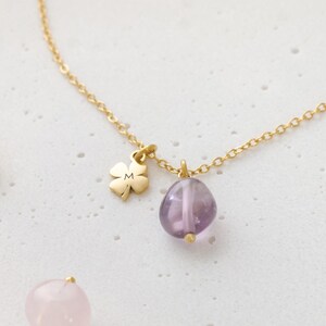 May include: A gold-colored necklace featuring a four-leaf clover charm with the letter "M" and a purple amethyst stone pendant. The necklace is displayed on a white surface.