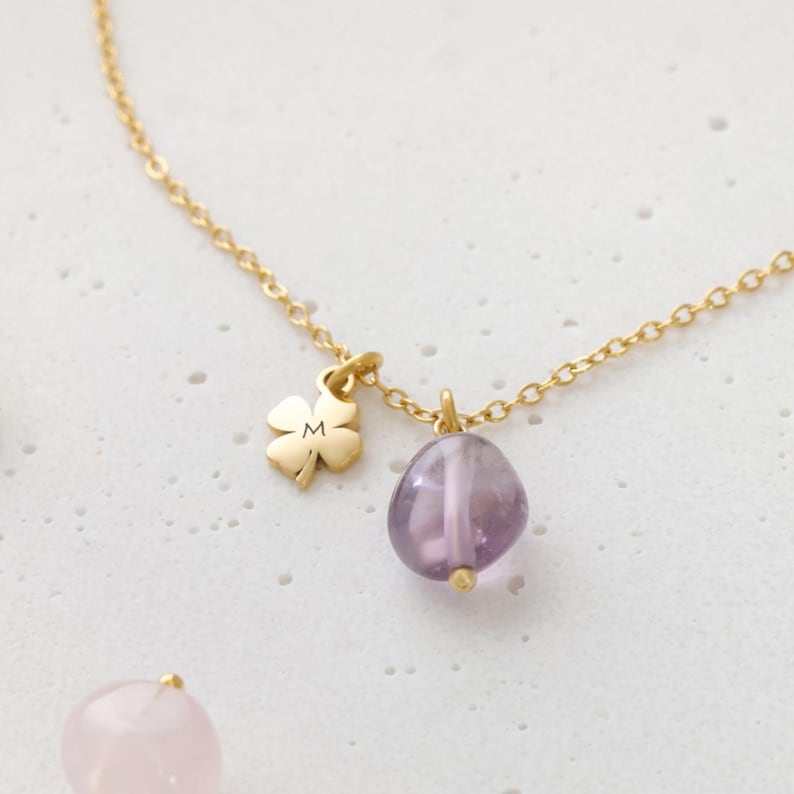 May include: A gold-colored necklace featuring a four-leaf clover charm with the letter "M" and a purple amethyst stone pendant. The necklace is displayed on a white surface.