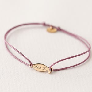 May include: A delicate pink cord bracelet with a gold charm engraved with the name "Lean" and a four-leaf clover.