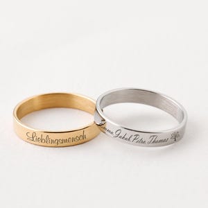 Könnte beinhalten: Zwei gravierte Ringe aus Edelstahl. Ein Ring ist golden und mit dem Text "Lieblingsmensch" graviert. Der andere Ring ist silber und mit dem Text "Emma. Jakob. Petra. Thomas" und einem kleinen Baum graviert.