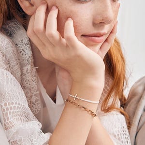 May include: A young person wearing a white lace top with a gold chain necklace and two bracelets on their wrist. One bracelet is a thin white string with a small gold cross charm. The other bracelet is a gold chain link bracelet.