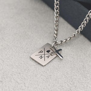 Könnte beinhalten: Silberfarbene Halskette mit einem rechteckigen Anhänger mit den Buchstaben P, N und A sowie einem Pfotenabdruck. Ein kleiner Kreuzanhänger hängt daneben. Die Kette ist im Curb-Stil gehalten.
