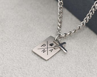 May include: Silver-toned necklace featuring a rectangular pendant with the letters P, N, and A, and a paw print. A small cross charm hangs next to the pendant. The chain is a curb style.