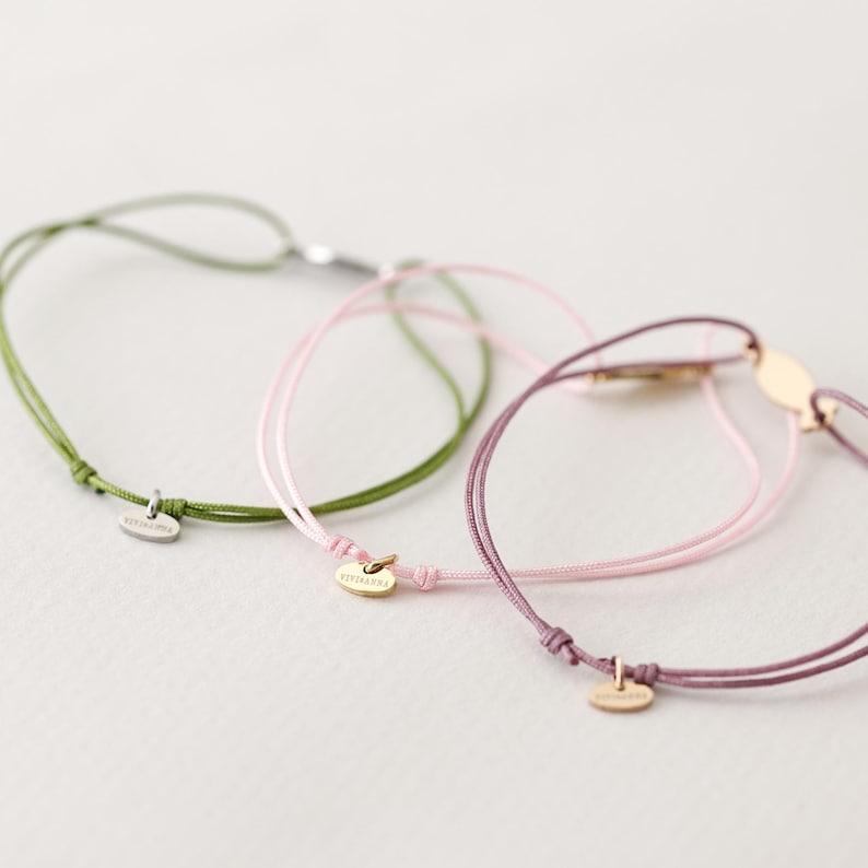 May include: Three friendship bracelets with different colored cords and small oval charms. The bracelets are green, pink, and purple.