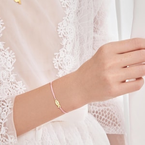 May include: A pink string bracelet with a small gold charm in the shape of a fish. The charm has the word "faith" engraved on it.