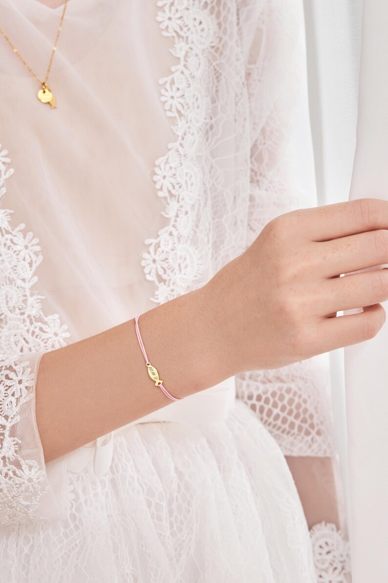May include: A pink string bracelet with a small gold charm in the shape of a fish. The charm has the word "faith" engraved on it.