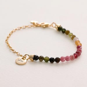 May include: Gold chain bracelet with a personalised charm that says "Lana" and a multi-coloured gemstone bead strand. The beads are green, black, pink, and yellow.