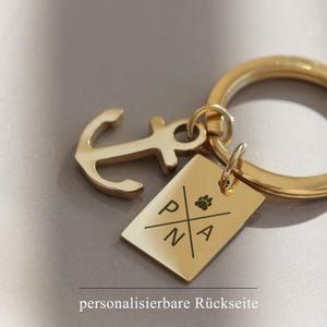 May include: Gold-toned keychain featuring an anchor charm and a rectangular tag engraved with the letters "PNA" and a paw print. The tag has a customisable back.