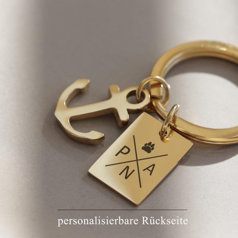 May include: Gold-toned keychain featuring an anchor charm and a rectangular tag engraved with the letters "PNA" and a paw print. The tag has a customisable back.